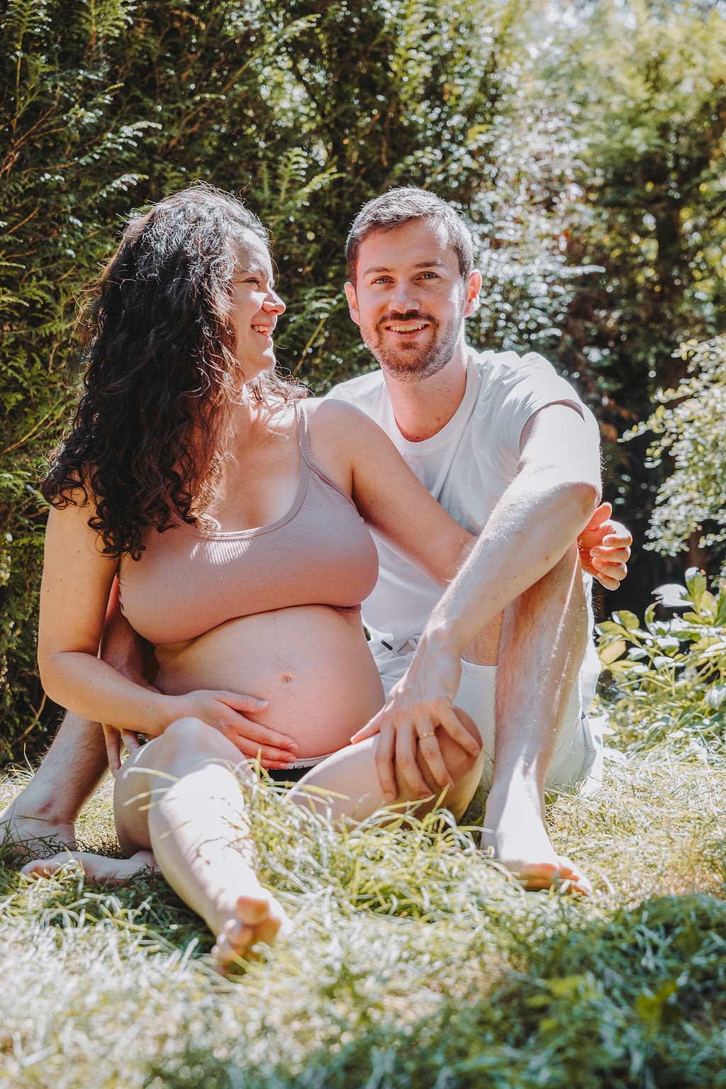 Couple assis dans l'herbe qui se tient dans les bras. La femme est enceinte et pose sa main sur son ventre avec le sourire