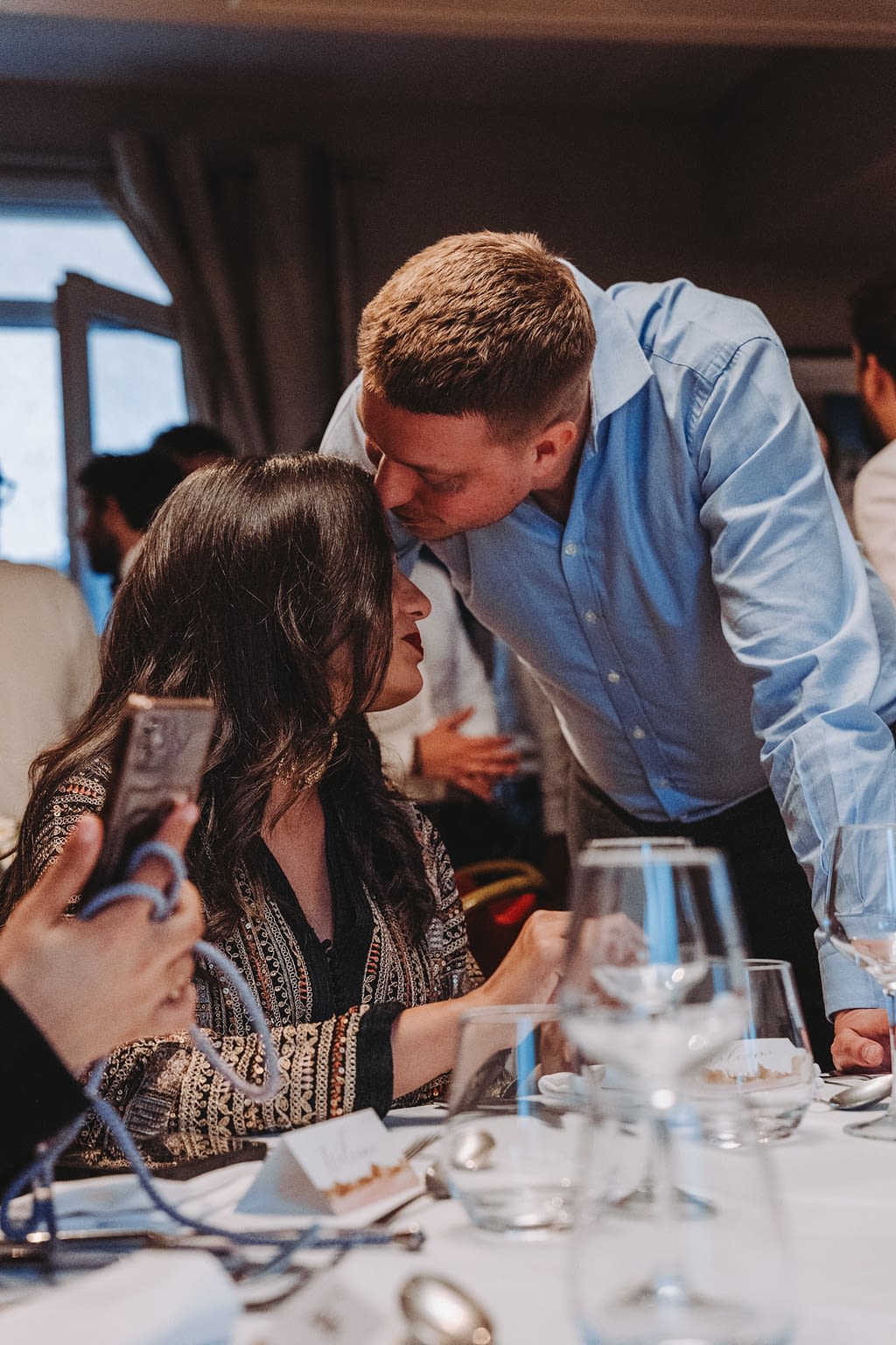 Photographie d'un couple de jeunes mariés pendant le dîner. Le marié fait un baiser sur le front à la mariée