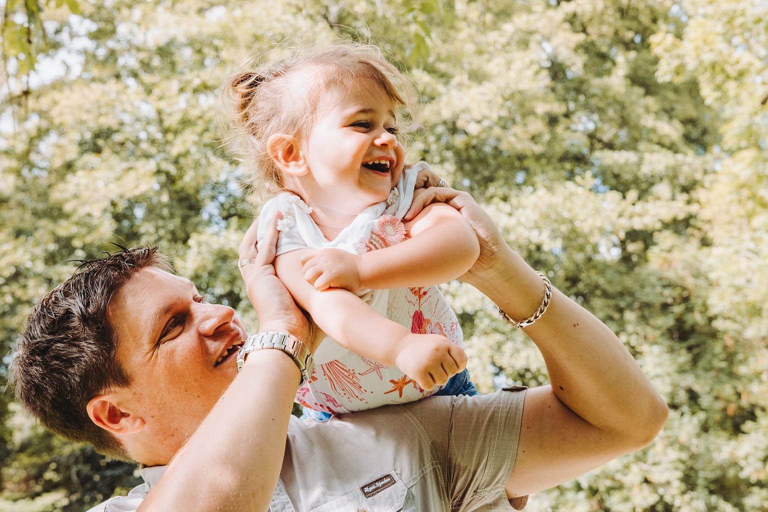 Photographie d'un papa et sa fille il la porte en l'air et joue avec elle dans un parc