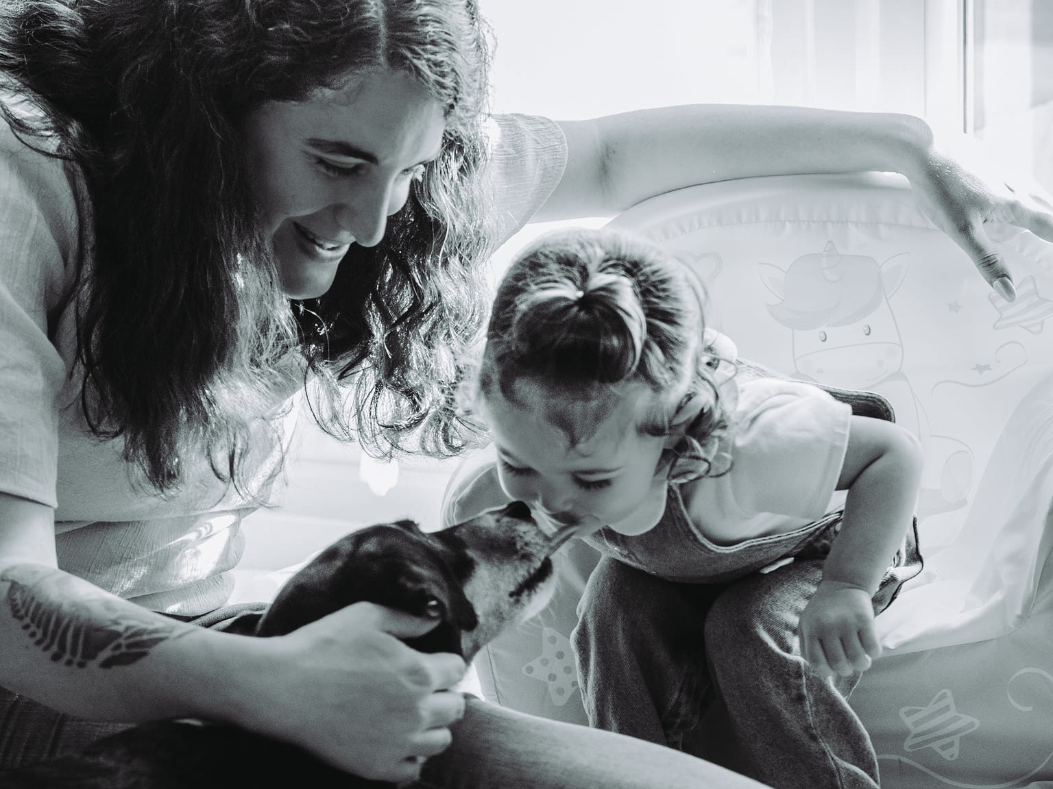 Photographie d'une maman avec sa fille et leur chien. Le chien lèche le bout du nez de la petite fille